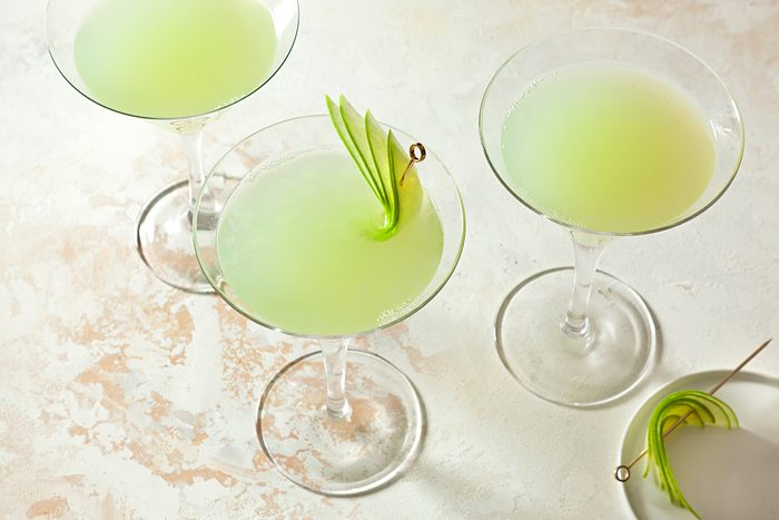 Three martini glasses filled with a light green cocktail are arranged on a textured surface. Each drink has a garnish of thinly sliced green apple skewered on a toothpick.