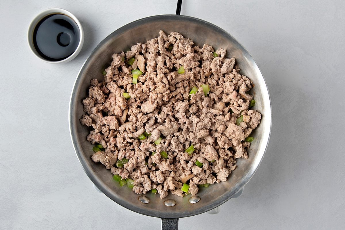 A pan filled with cooked ground turkey mixed with green pepper pieces. A small bowl containing a dark sauce is placed on the side, on a light gray surface.