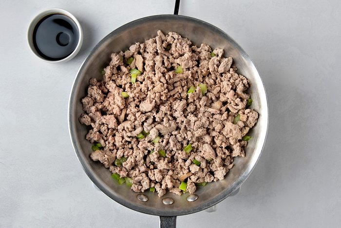 A pan filled with cooked ground turkey mixed with green pepper pieces. A small bowl containing a dark sauce is placed on the side, on a light gray surface.