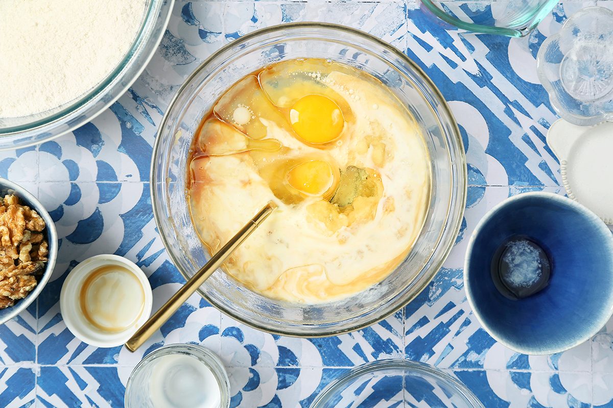 A bowl with eggs, milk, and vanilla essence on a blue patterned surface, ready for mixing. Surrounding the bowl are small bowls containing walnuts, flour, and various ingredients for baking.
