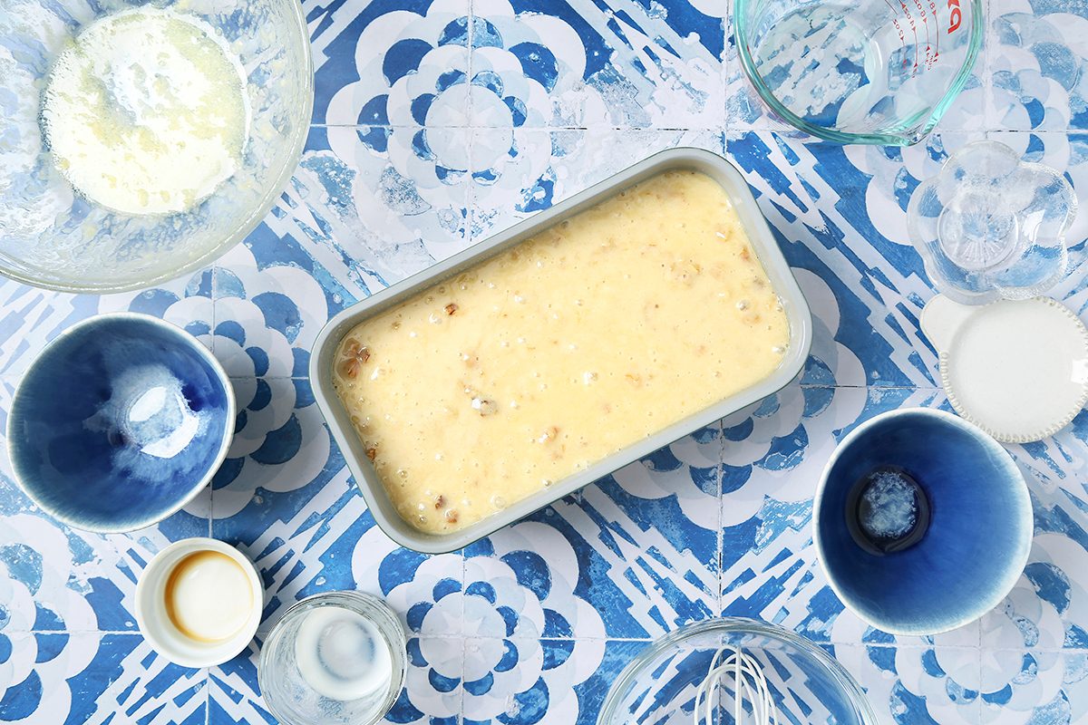 A loaf pan filled with batter sits on a blue and white patterned surface, surrounded by empty blue bowls, a glass measuring cup, and remnants of ingredients such as flour and milk.