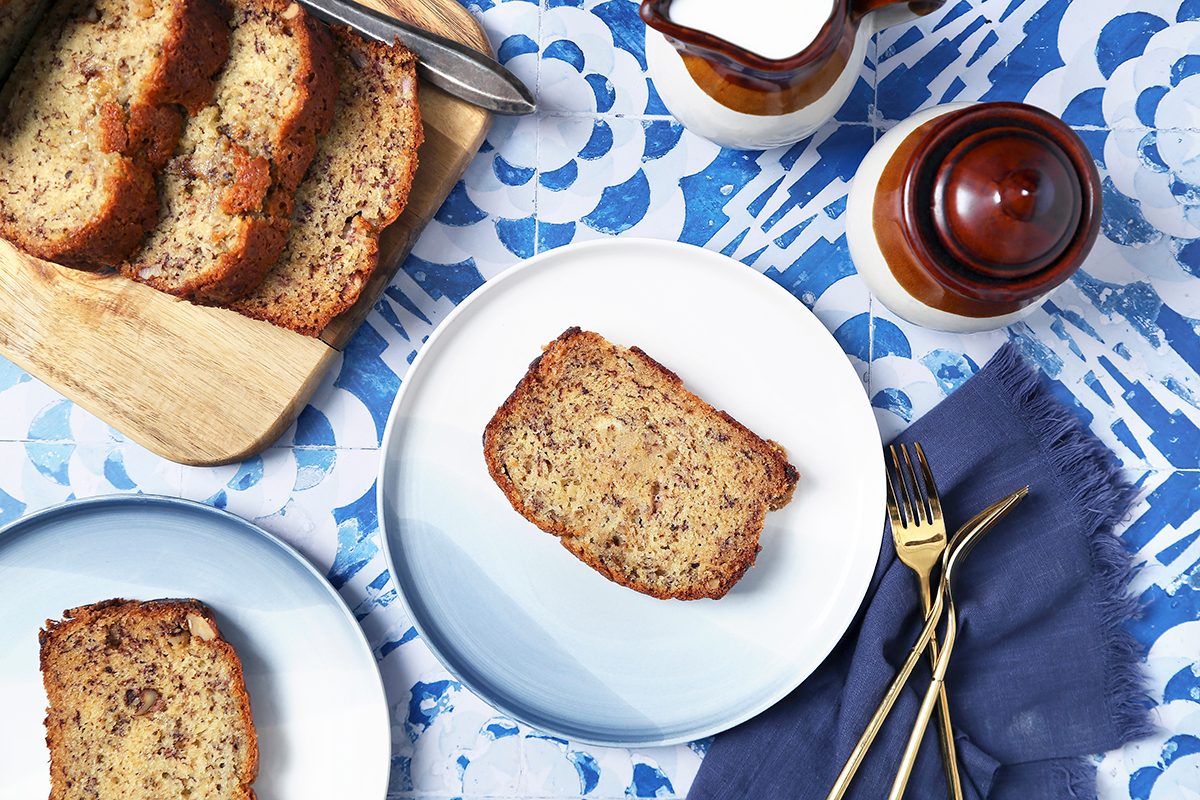 Slices of banana bread on a wooden board and a blue and white plate. They sit on a blue-patterned tablecloth beside a dark blue napkin with gold forks and knives, and ceramic containers, one holding milk.