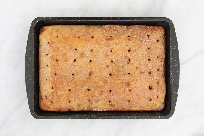 A rectangular cake with holes on top sits in a black baking tray. The cake appears lightly browned and evenly baked, placed on a white marble surface.
