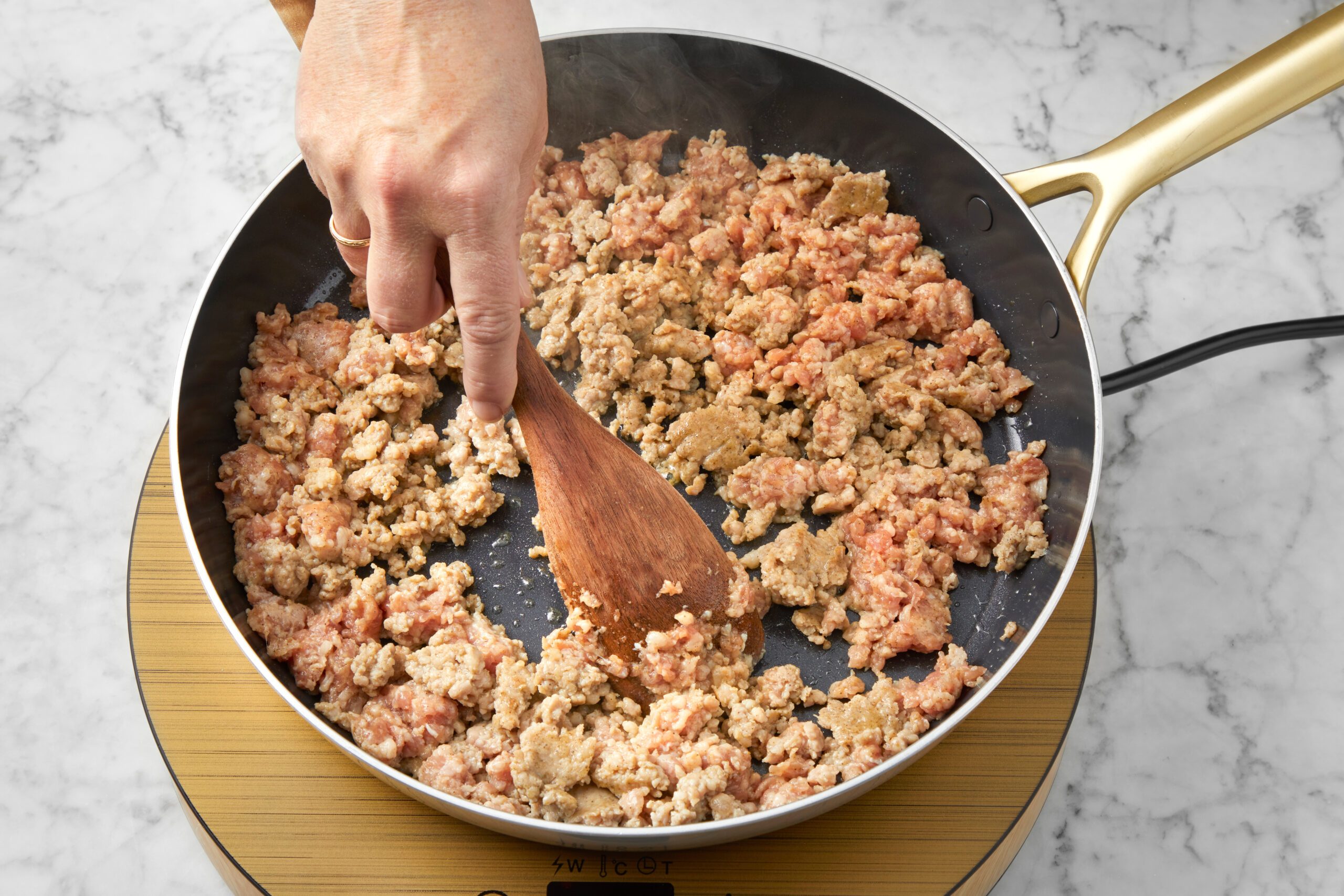 A hand stirs ground meat in a non-stick frying pan using a wooden spatula. The pan is placed on a circular hot plate over a marble countertop. The meat is partially cooked, showing a mix of brown and pink tones.