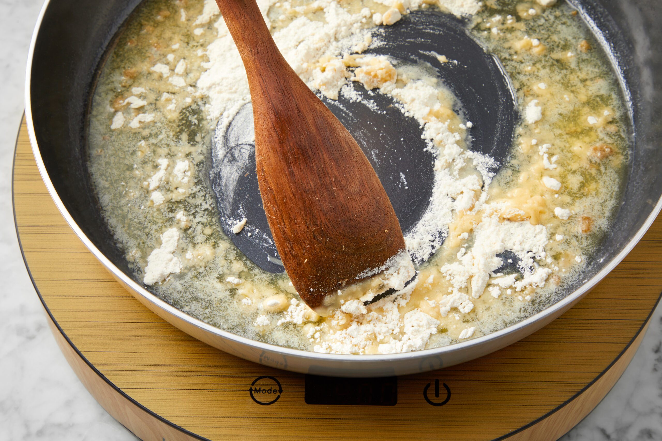 A wooden spoon stirs flour and butter in a pan on a stove, creating a roux. The mixture is in the early stages, with some clumps and a light color, over a digital induction cooktop with a wooden finish.