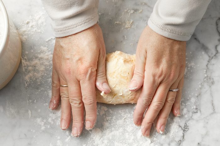 Hands kneading dough on a floured marble surface. The person is wearing a light-colored long-sleeve shirt and rings on both hands. A bowl is partially visible on the left side.