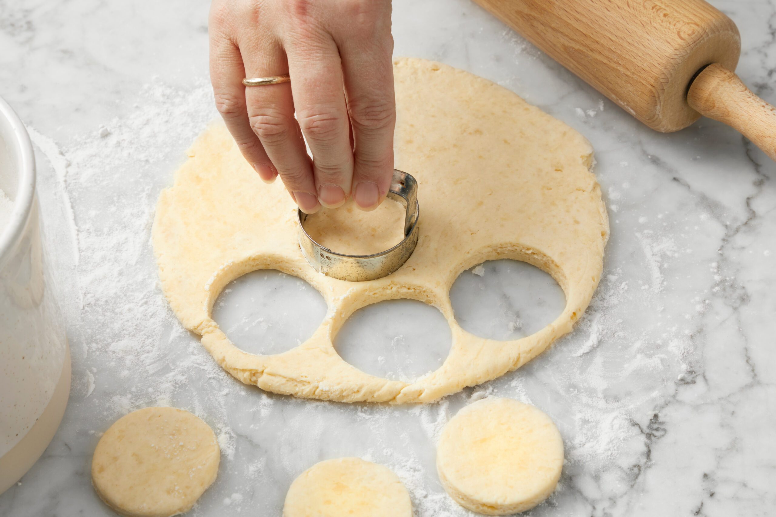 A hand uses a round cookie cutter to make circles in rolled-out dough on a floured marble surface. A wooden rolling pin is visible in the top right corner, and cut dough rounds are placed nearby.