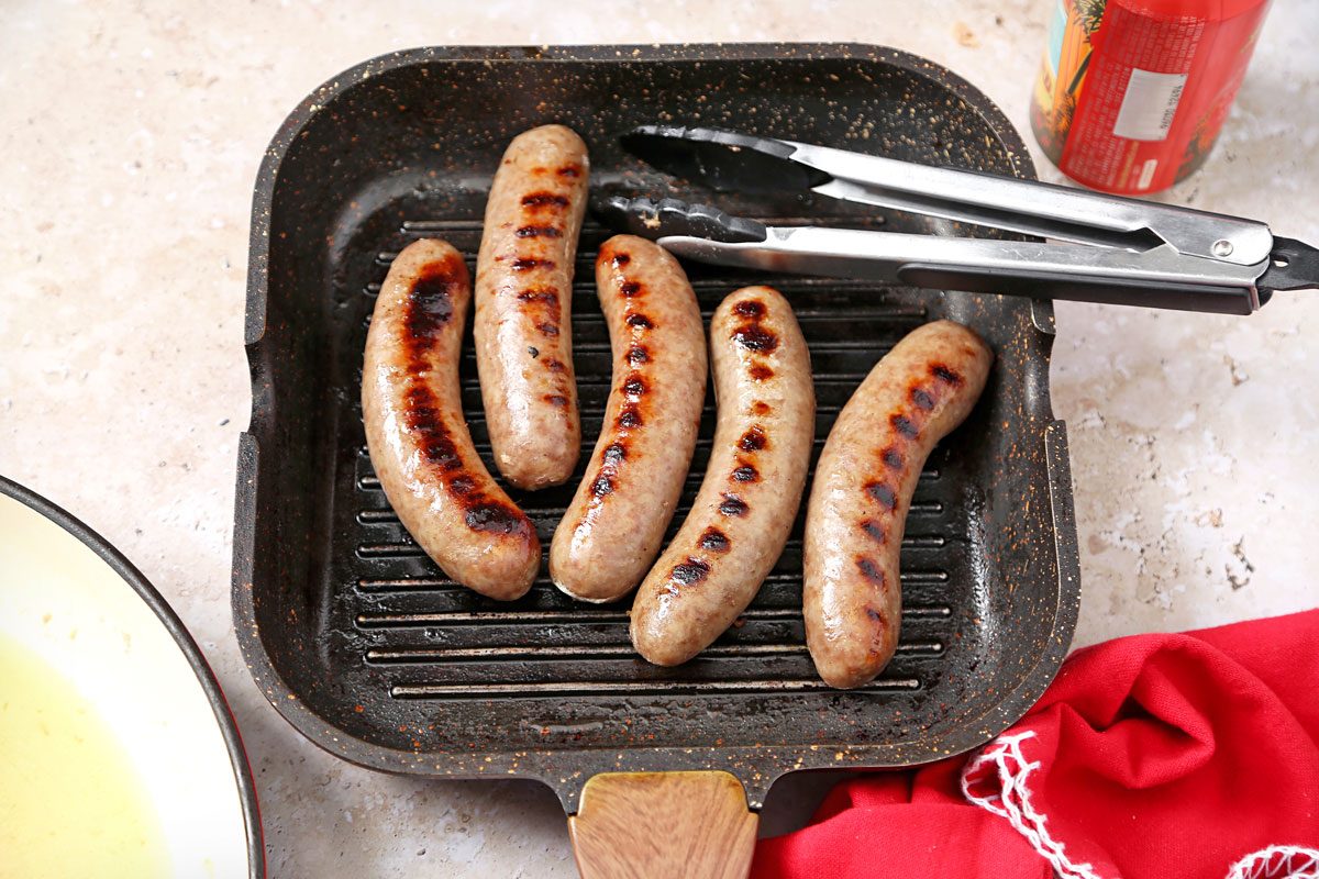 overhead shot of a black grill pan with a wooden handle, filled with five grilled sausages, a pair of tongs, a red and white checkered cloth in the lower right corner, and an empty white pan to the left of the grill pan