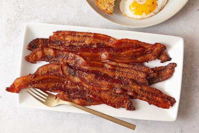 A rectangular white plate with crispy cooked bacon strips and a gold fork on the side. In the background, there's a partially visible plate with a sunny-side-up egg and toast. The setting is on a light-colored surface.