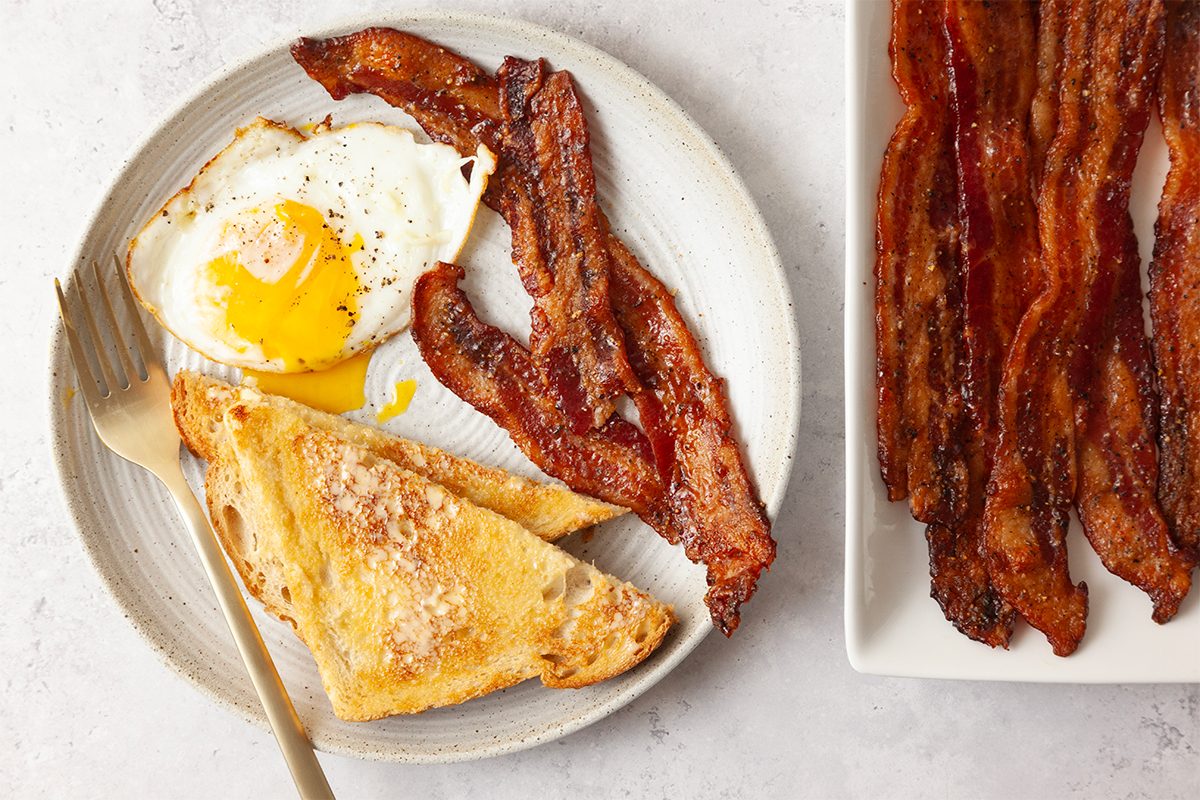 A breakfast plate with a sunny-side-up egg, crispy bacon strips, and a slice of buttered toast. Beside it, more bacon strips are placed on a rectangular white dish. A fork rests on the plate.