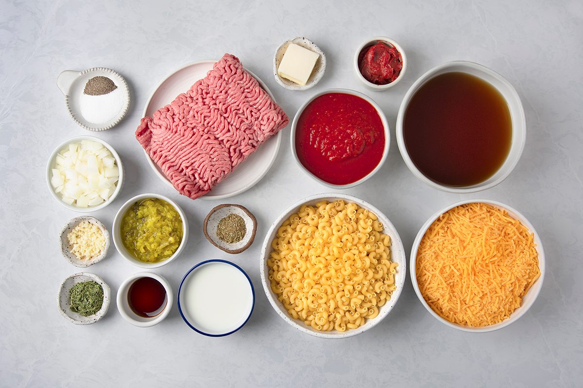 Various ingredients are arranged on a light surface, including ground meat, pasta, grated cheese, diced onions, tomato paste, broth, seasonings, butter, cream, and spices in small bowls.