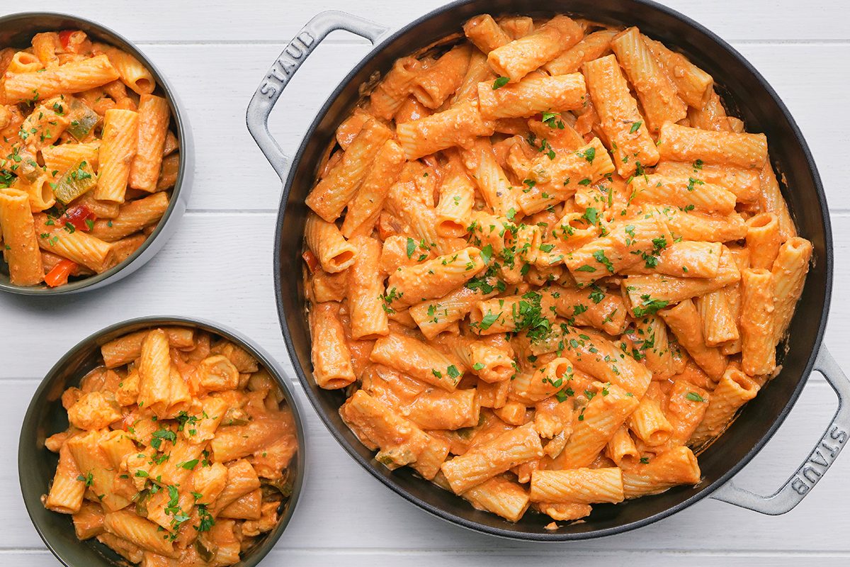 A large pan of creamy tomato rigatoni is garnished with chopped parsley. Two smaller bowls hold portions of the same pasta on a white wooden table.
