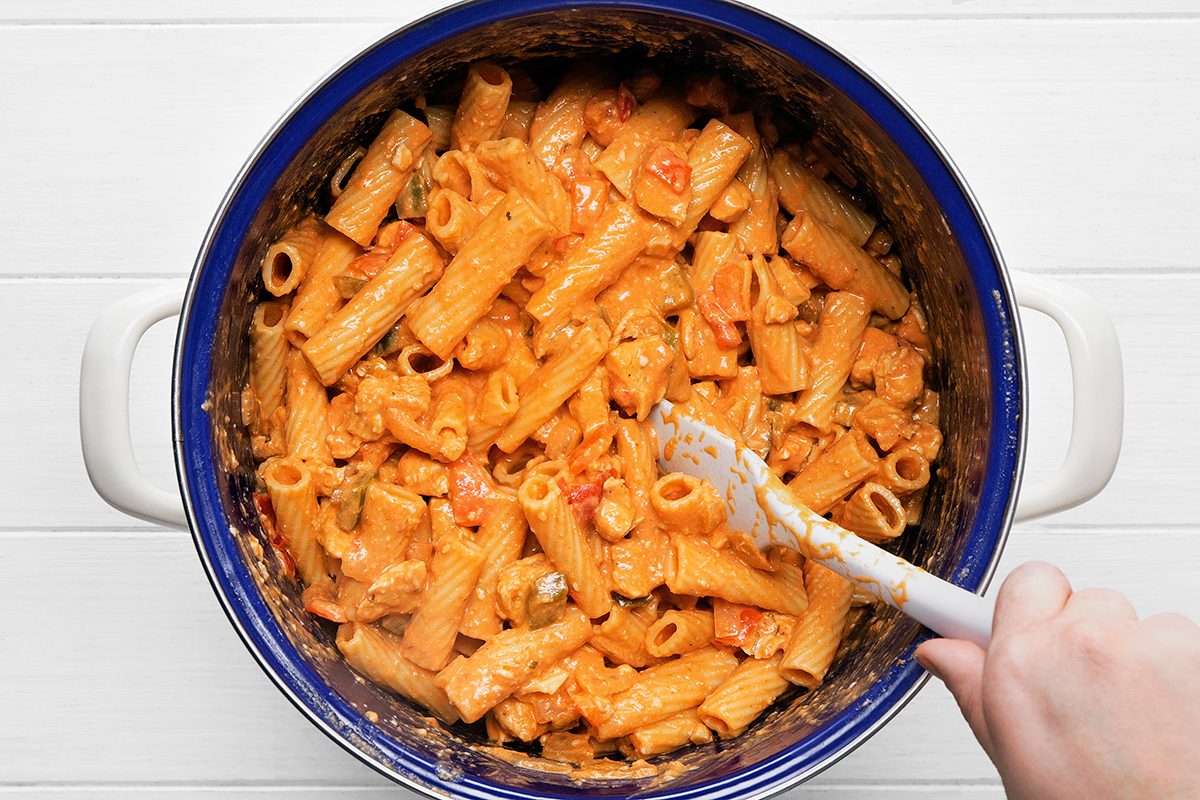A person stirs creamy tomato pasta in a large blue and white pot. The pasta is rigatoni and is coated in a thick orange sauce with pieces of tomato and herbs visible. The pot rests on a white surface.