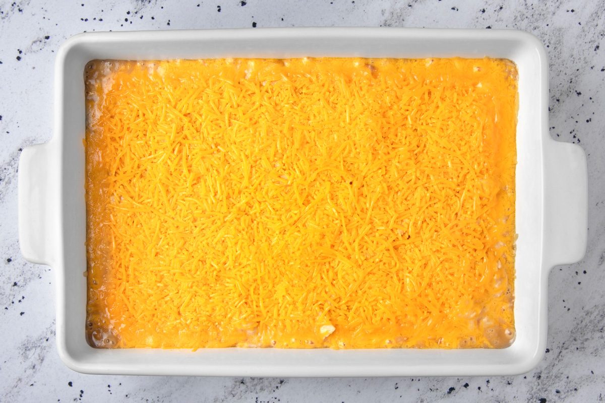 overhead shot of a white ceramic baking dish filled with a layer of shredded cheese; the cheese is bright yellow and covers the entire surface of the dish; the dish is sitting on a white surface with gray speckles
