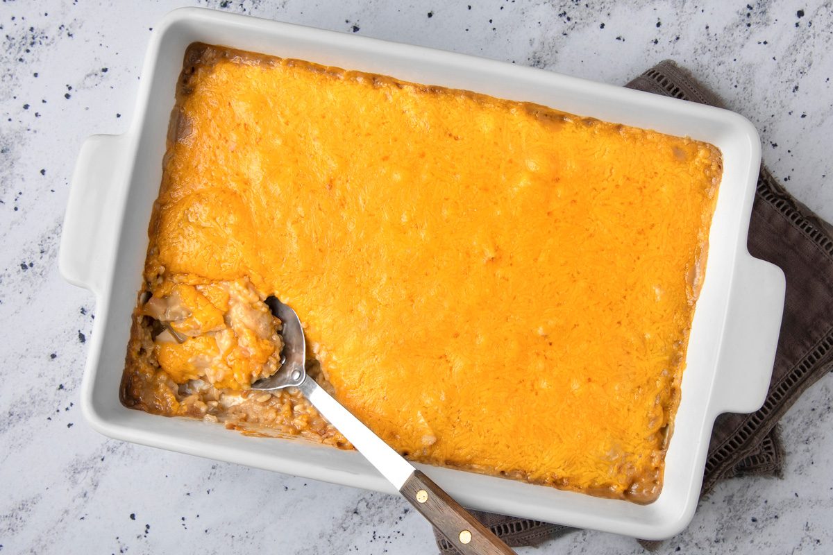 overhead shot of a white rectangular casserole dish filled with a cheesy dish, a portion of which has been scooped out with a silver spoon; the spoon has a wooden handle and is resting on a brown dish towel that is partially visible in the lower right corner of the image; the dish is resting on a white countertop