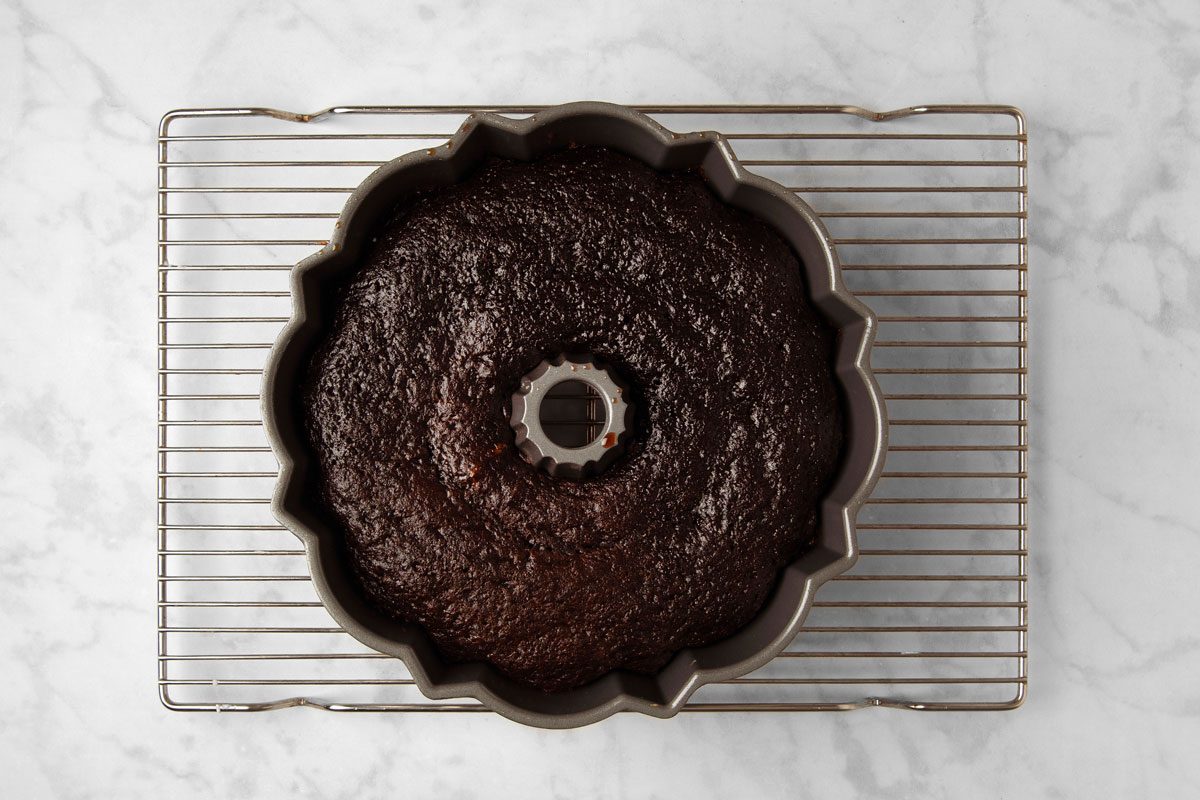 overhead shot of a baked chocolate cake in a bundt pan, cooling on a wire rack; the cake has a rich brown color and is covered in a thin layer of glaze; the cake is cooling on a wire rack that is placed on a marble countertop