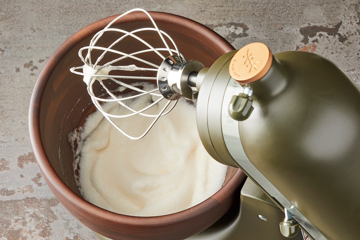 A green stand mixer with a metal whisk attachment is mixing a pale batter in a brown mixing bowl. The mixer is on a speckled countertop.