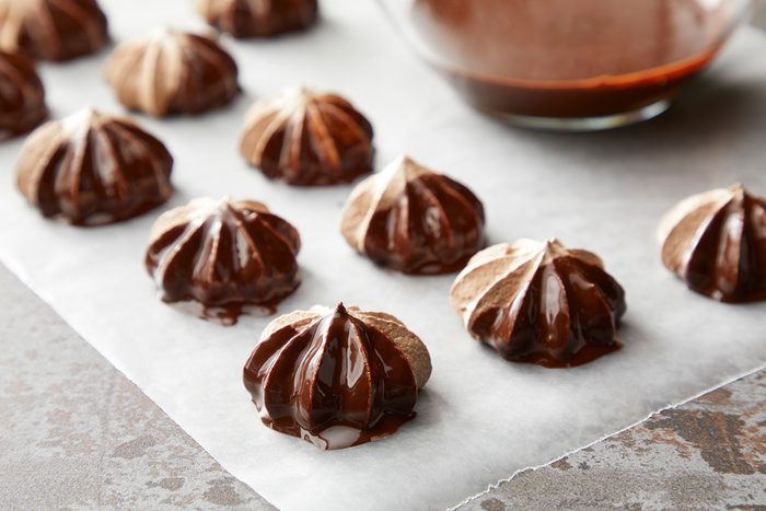 Chocolate meringue cookies on parchment paper, each dipped in melted chocolate. A bowl of chocolate sauce is visible in the background. The cookies have a swirled texture and are arranged in neat rows.