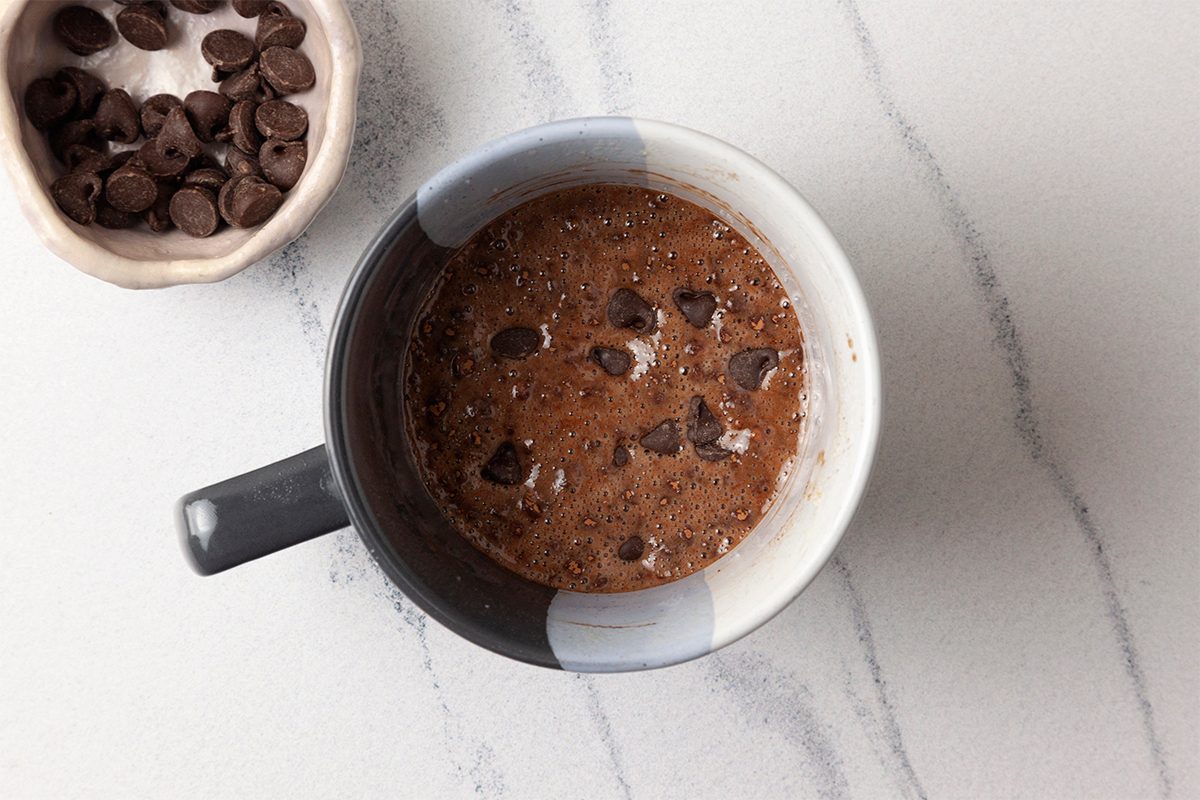 A mug filled with chocolate cake batter featuring chocolate chips on a marble countertop. There is a small bowl with chocolate chips next to the mug.