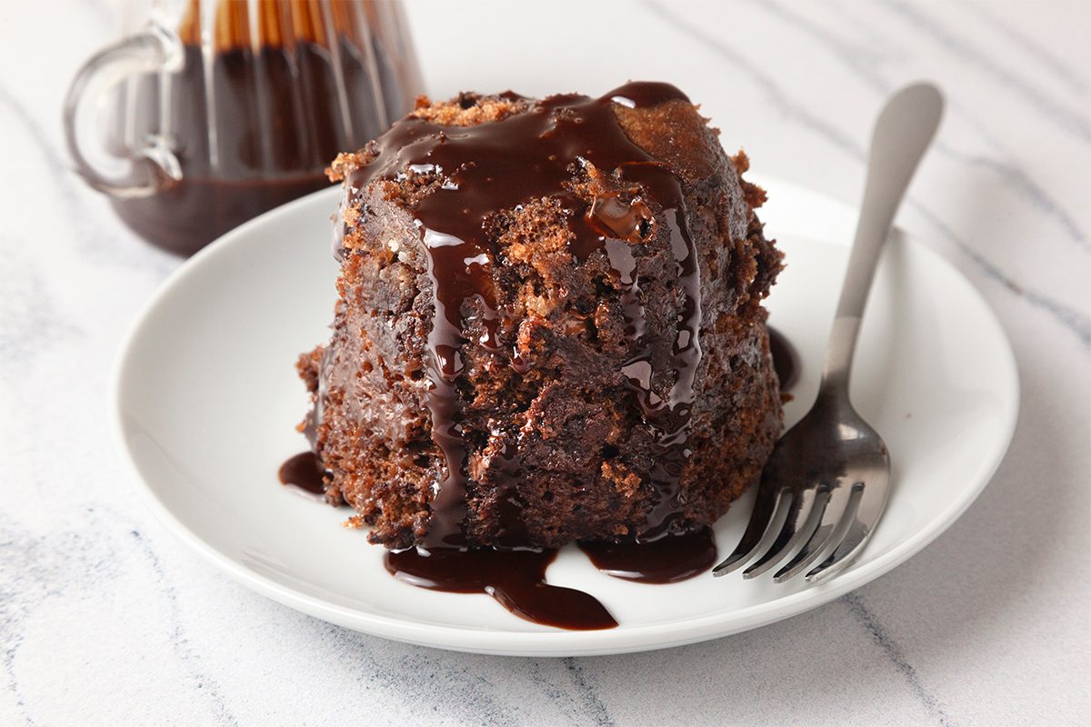 A chocolate lava cake sits on a white plate, topped with rich chocolate sauce that drips down the sides. A fork rests beside it. A glass jug filled with additional chocolate sauce is in the background on a marble surface.