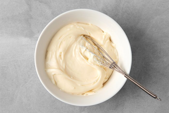 A white bowl filled with smooth, creamy batter. A metal whisk rests inside the bowl, partially covered by the batter. The bowl is placed on a grey textured surface.