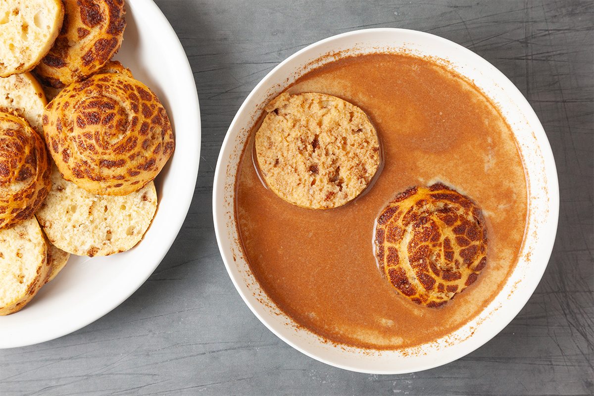 A bowl of rich, brown soup with two pieces of bread soaking in it. Beside the bowl, a plate holds several round, toasted rolls with a patterned crust, likely tiger bread. The background is a gray surface.