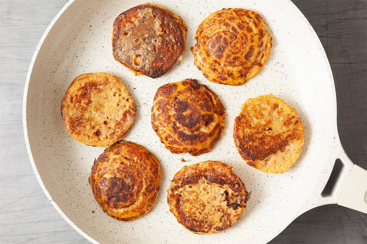 A white frying pan with seven round, golden-brown pancakes resting on its surface, each with slightly varied patterns and shades. The pan is on a gray background.