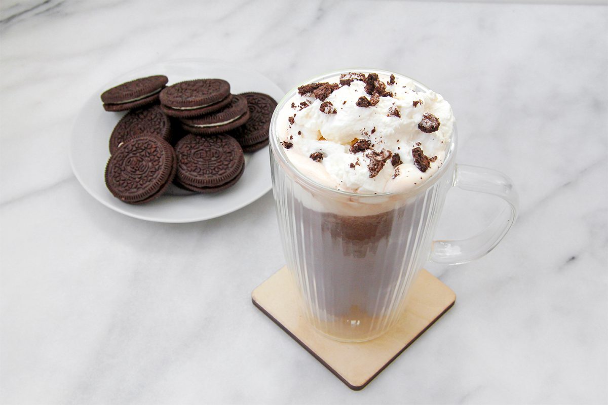 A glass mug of hot chocolate topped with whipped cream and chocolate crumbles sits on a coaster. Next to it is a plate with a stack of chocolate sandwich cookies on a white marble surface.