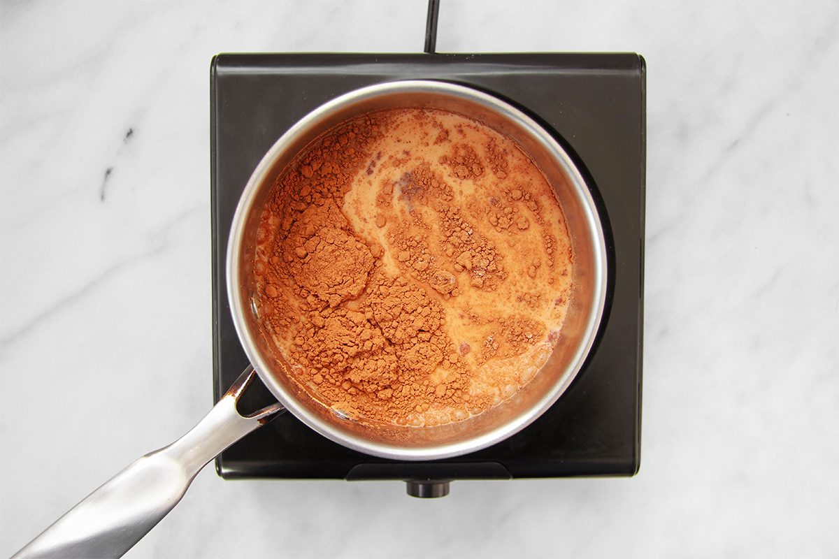 A saucepan on a black stovetop contains a bubbling chocolate mixture. The contents appear to be heating or cooking, with some frothy bubbles on the surface. The stovetop is placed on a white marble surface.
