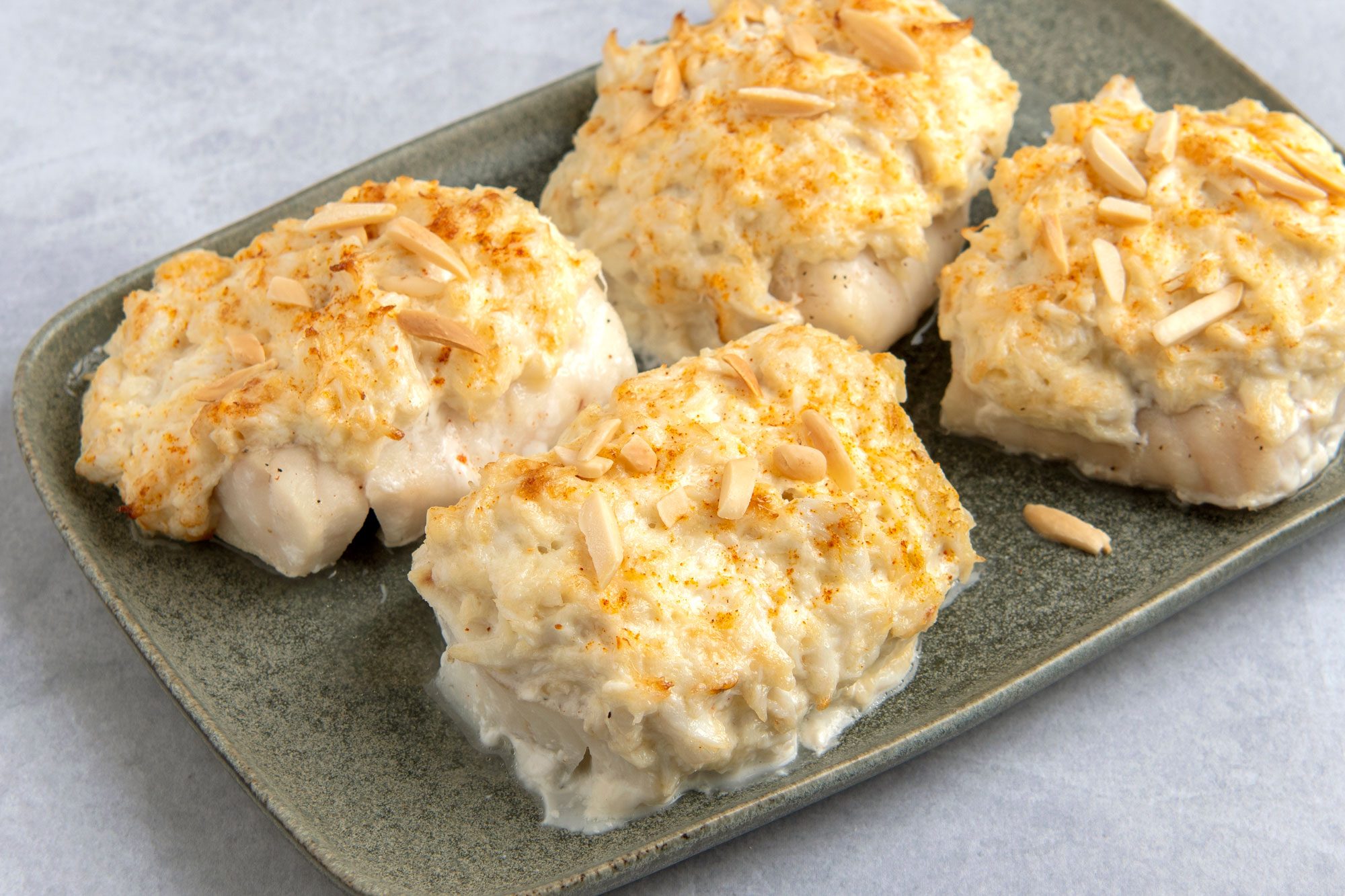 Four pieces of Crab-Topped Fish on a green plate and each piece is topped with a creamy sauce and sliced almonds