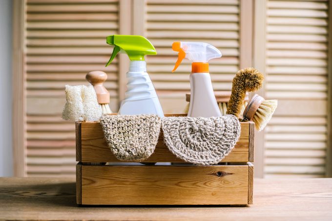 A wooden box filled with eco-friendly cleaning supplies, including spray bottles, scrub brushes, and cloths, is placed on a wooden surface. The background features light-colored, slatted shutters.