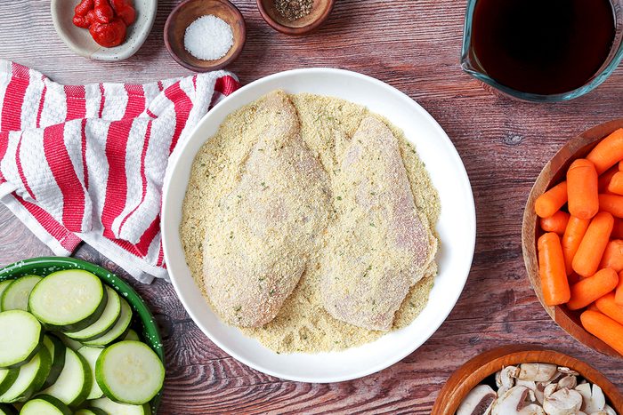Two breaded chicken fillets on a white plate surrounded by sliced zucchini, baby carrots, mushrooms, sauce, salt, and pepper. A red striped cloth is visible on a wooden surface.