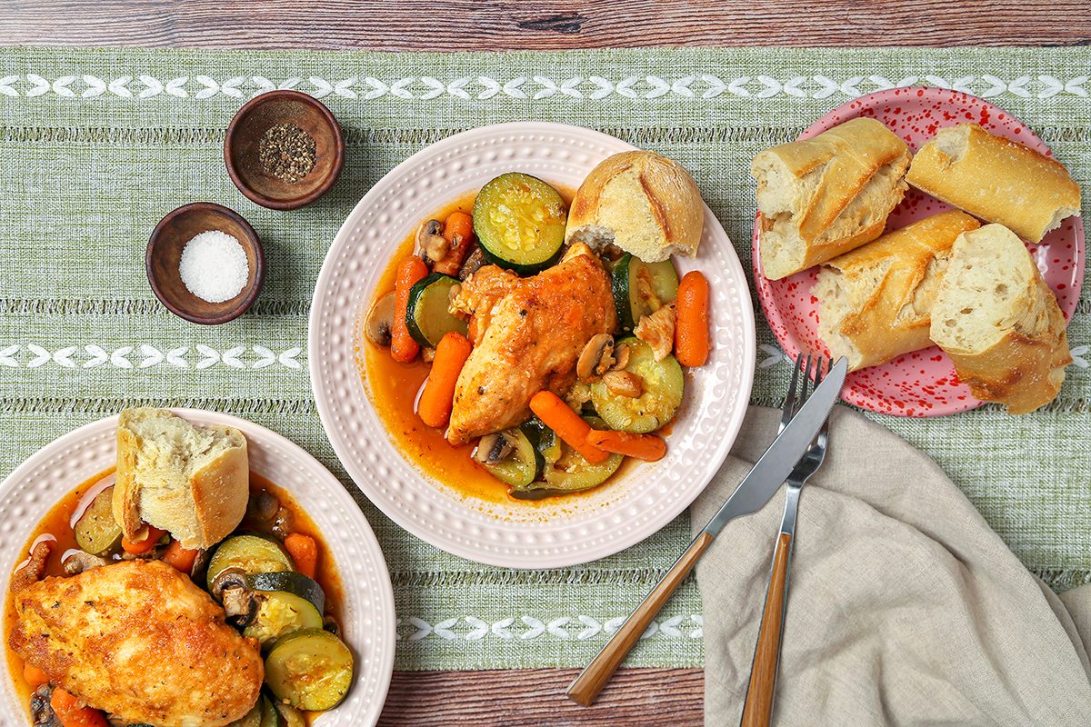 Plates of chicken with carrots, zucchini, and mushrooms in a sauce, served with bread. Additional bread is on a separate plate alongside. Utensils are placed on a napkin, and small bowls of pepper and salt are beside the dishes on a textured placemat.
