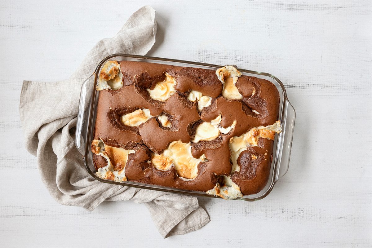 A glass baking dish filled with chocolate brownies topped with swirls of melted marshmallows. A beige cloth napkin is placed beside the dish on a white wooden surface.