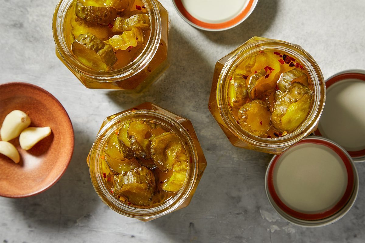 Three jars filled with pickled cucumbers are arranged on a gray surface. Nearby, a small dish holds garlic cloves, and two lids rest beside the jars.