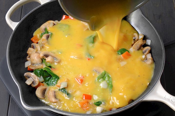 Egg mixture being poured into a skillet with mushrooms, spinach, chopped onions, and red bell peppers.