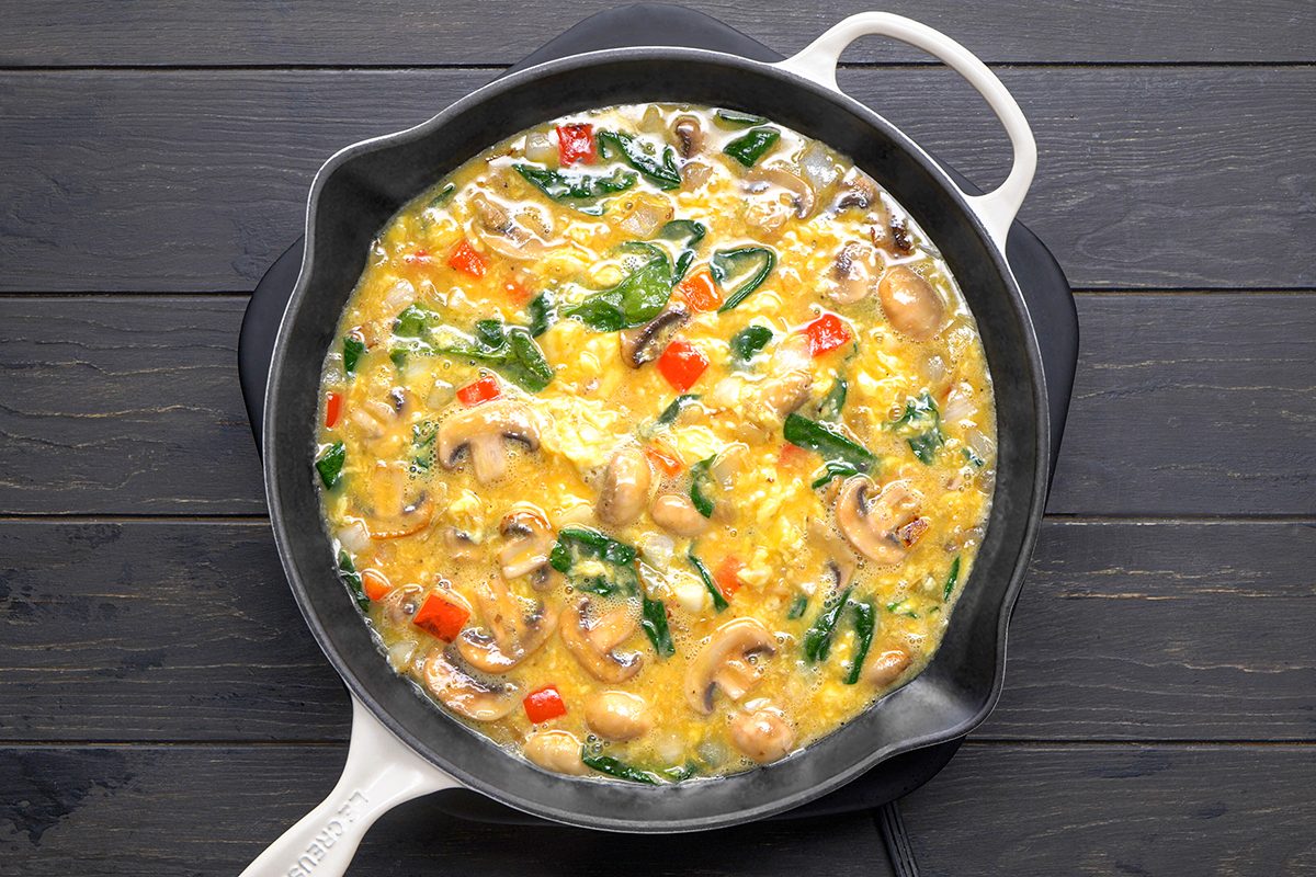 A skillet filled with an omelet on a dark wooden table. The omelet contains mushrooms, spinach, diced red bell peppers, and a blend of cheeses, giving it a colorful appearance. The skillet has a white handle.