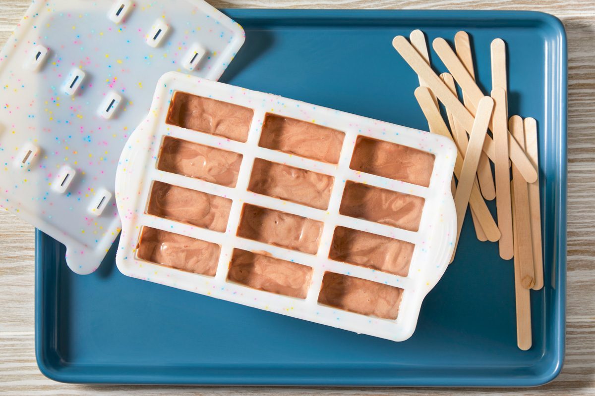 overhead shot of a blue rectangular dish with a white ice pop mold filled with chocolate fudge batter; on the right side of the dish are a bunch of wooden popsicle sticks and a single popsicle stick lying on the side; the white ice pop mold is covered with colorful sprinkles; the dish is on a wooden surface