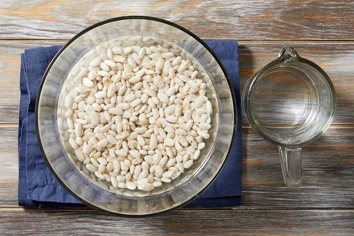 A glass bowl filled with white beans soaking in water sits on a wooden surface with a blue cloth underneath. Next to it, there is an empty glass measuring cup.