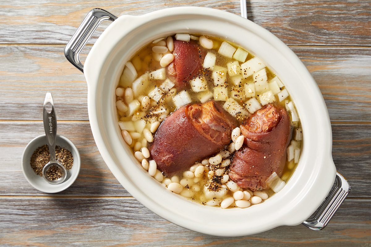 A crockpot filled with chopped onions, white beans, and smoked ham hocks in broth, seasoned with pepper. A small bowl with a spoon containing more seasoning sits beside it on a wooden surface.