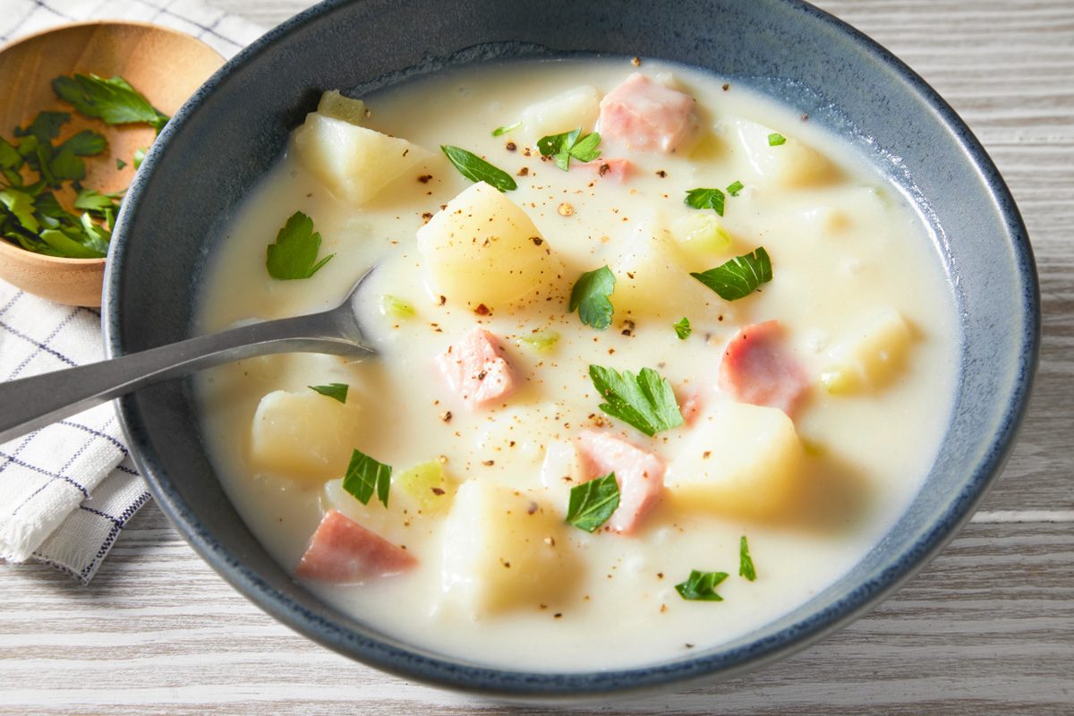 3/4th shot of A bowl of Ham and Potato Soup, a few sprigs of parsley are scattered throughout; the bowl is sitting on a light wood table, there is a small bowl of chopped parsley and a cloth napkin to the left of the soup bowl