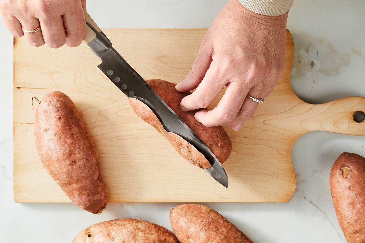 A person cutting sweet potatoes on a wooden cutting board with a large knife. The board is on a light-colored countertop, and several uncut sweet potatoes are placed around the cutting board.