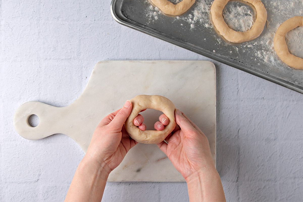 Hands shaping a dough ring on a marble cutting board. A metal baking tray with more dough rings sprinkled with flour is in the background.