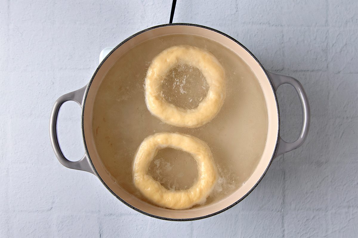 A pot of boiling water with two round dough pieces being cooked inside. The pot has two handles and is placed on a light-colored surface.