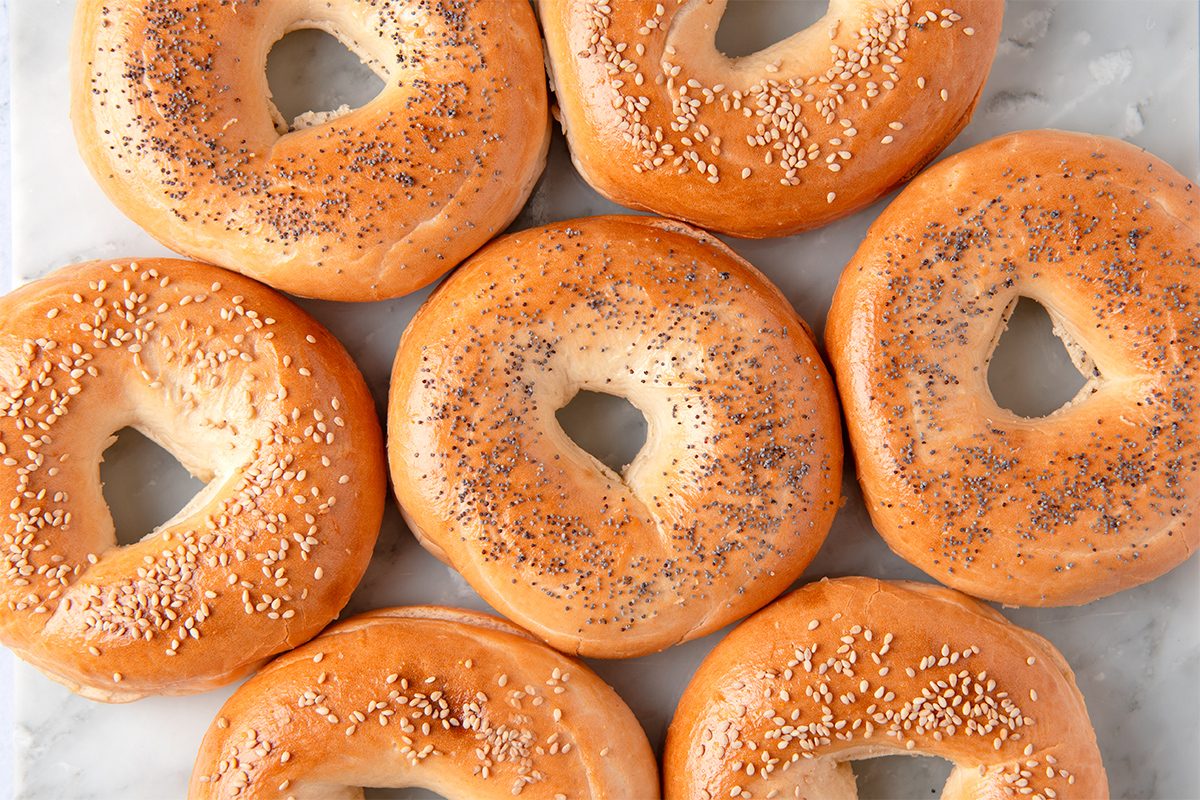 Seven bagels topped with poppy seeds and sesame seeds are arranged on a marble surface. They're golden-brown and evenly spaced, creating a visually appealing pattern.