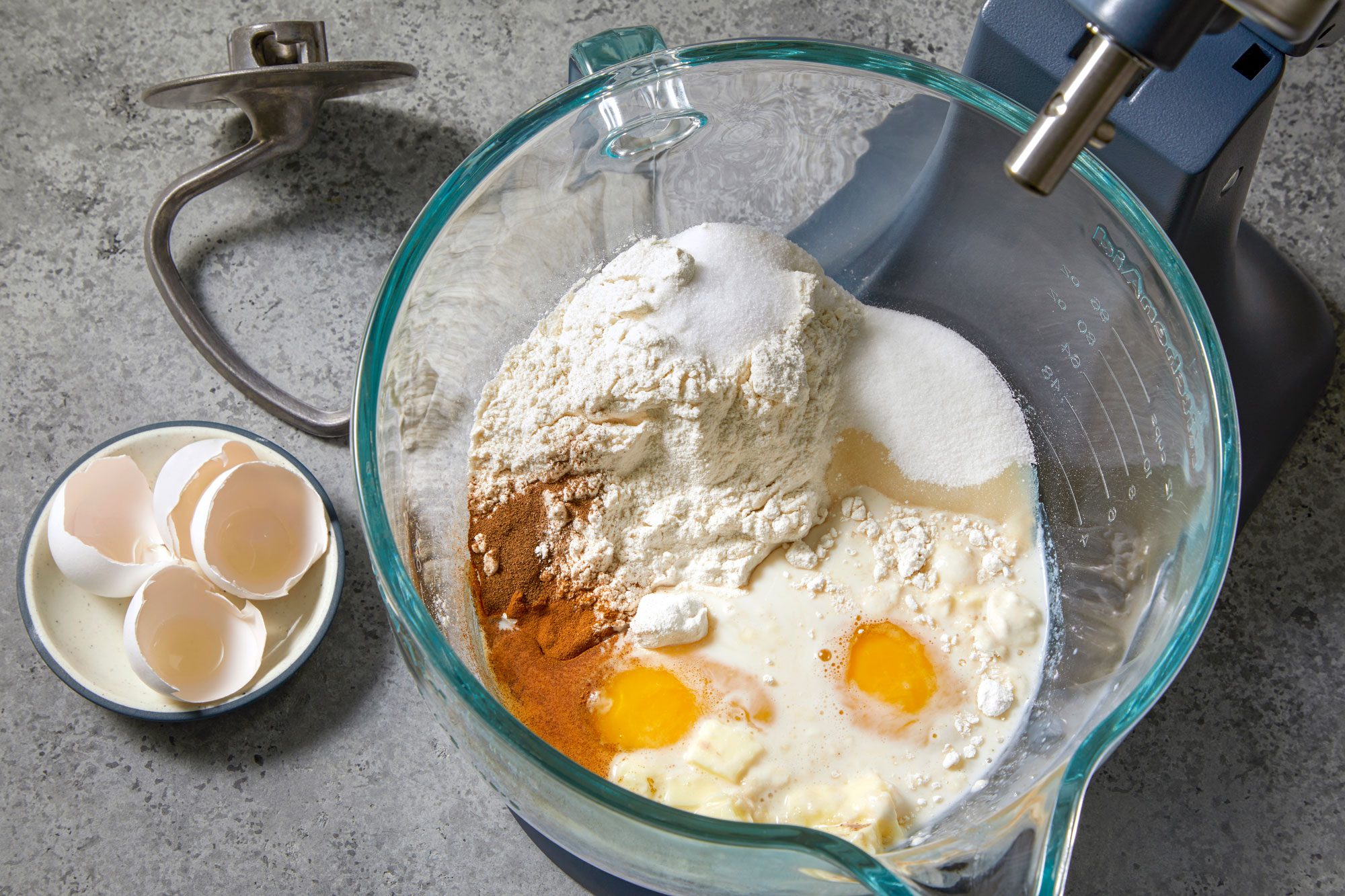 overhead shot of a glass bowl of a stand mixer contains the ingredients for a recipe; some cracked eggshells are in a small white bowl with a blue rim; the eggshells are sitting on the counter next to the bowl of ingredients