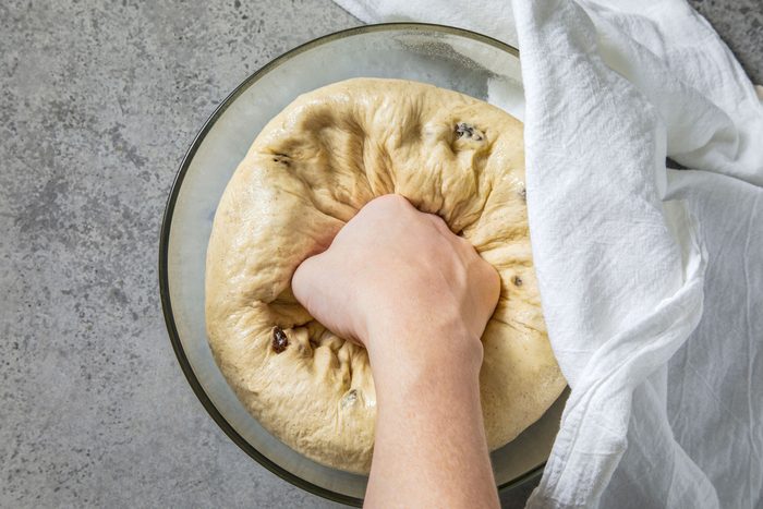 overhead shot of a hand pressing down on a dough ball in a glass bowl; the dough is sitting on top of a white cloth that is draped over the side of the bowl; the background is a light gray countertop