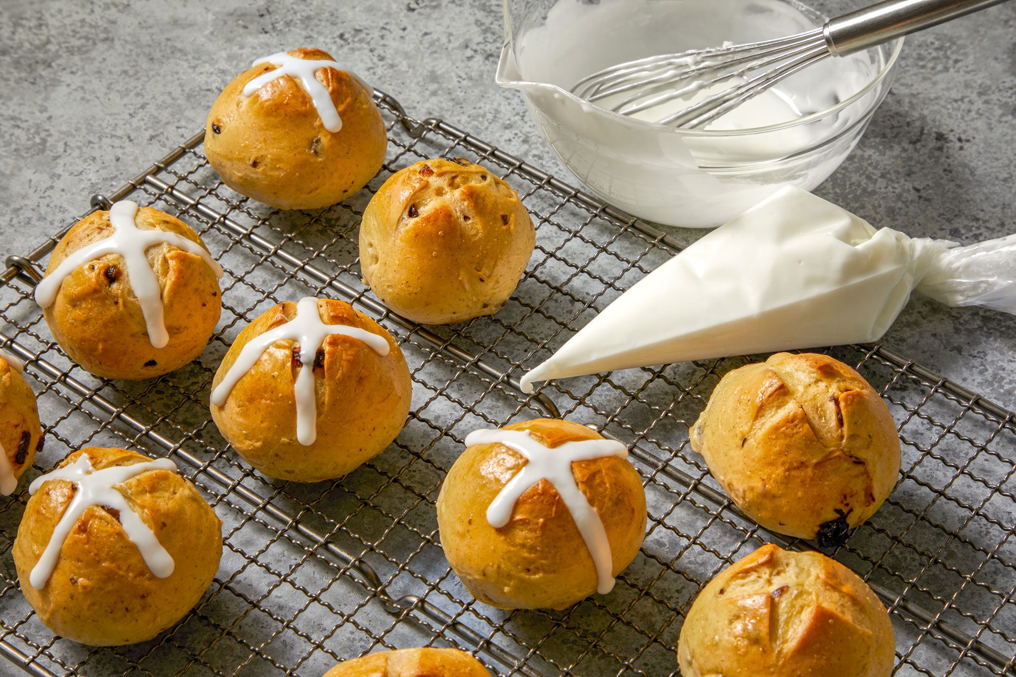 3/4th shot of a plate of Traditional Hot Cross Buns; the buns are arranged in a criss-cross pattern on a wire rack, with a few buns in the foreground and a few in the background; the buns are surrounded by a bowl of white icing, a piping bag of white icing, and a whisk
