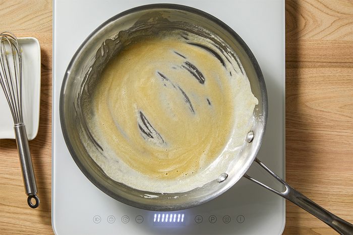 A stainless steel pan on a white induction cooktop contains a light brown roux being stirred. A metal whisk is placed nearby on the wooden countertop.