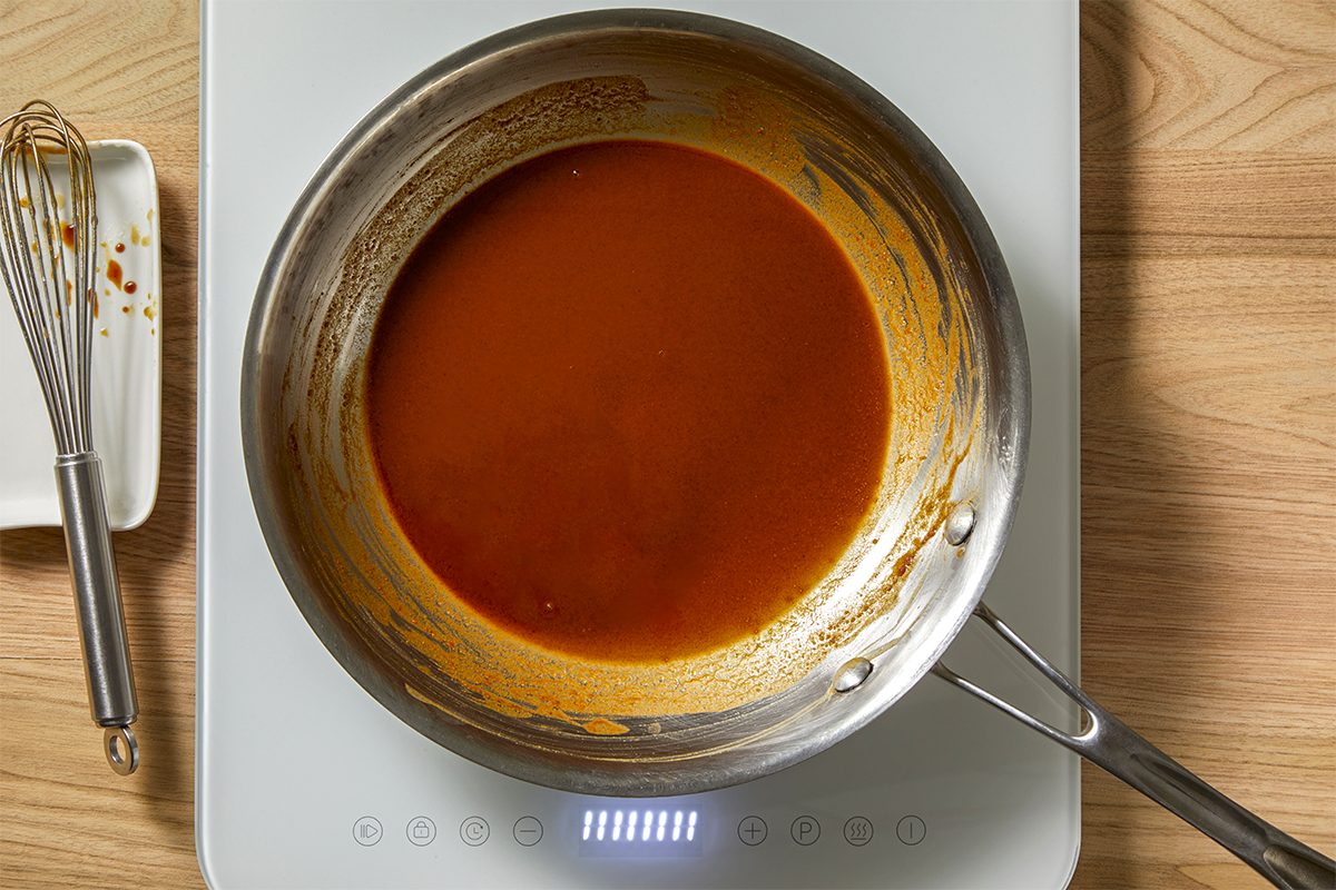 A stainless steel pan with brown butter sauce sits on an electric stove. A whisk rests on a dish nearby, both on a wooden surface. The sauce is smooth and golden-brown, partially coating the sides of the pan.